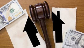 A judge's gavel is placed over a torn piece of paper representing a house and stacks of $100 bills.