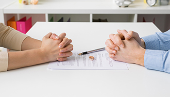 A man and a woman sit across from each other at a table, their hands clasped over a divorce document.