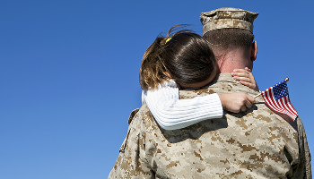 A soldier in a camouflage uniform hugs a young girl, who holds a small American flag over his shoulder.