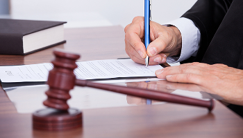 A judge in black robes writes with a pen on a legal document with a wooden gavel in the foreground.