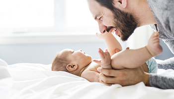A smiling bearded man looks down at a baby lying on a bed.